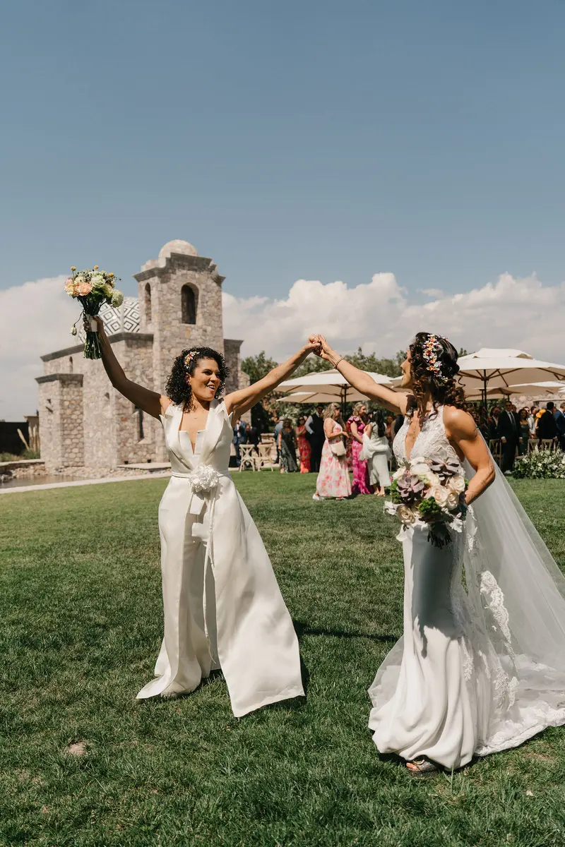 The two brides raising their bouquets in celebration after the ceremony, chapel tower behind them at Hacienda Los Arcángeles