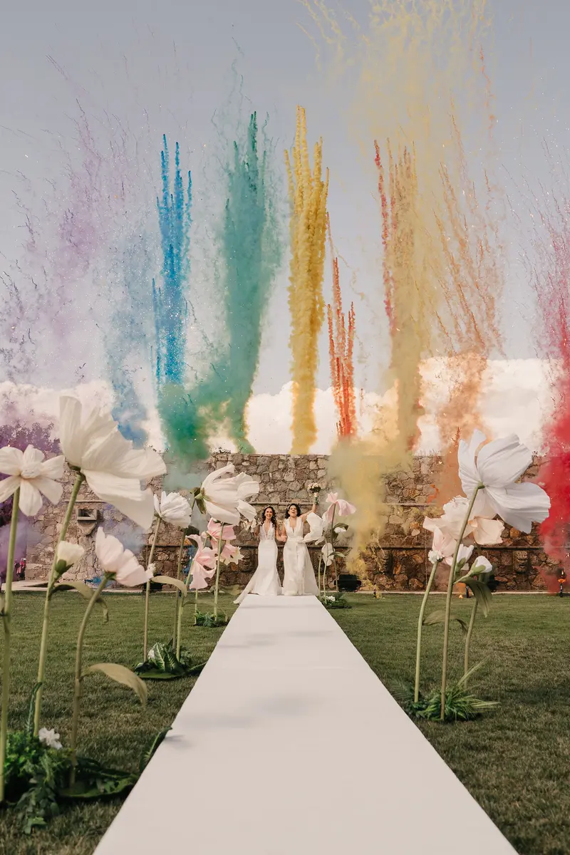 The two brides walking into the party under a burst of colored smoke, flanked by an aisle of giant paper flowers at Hacienda Los Arcángeles