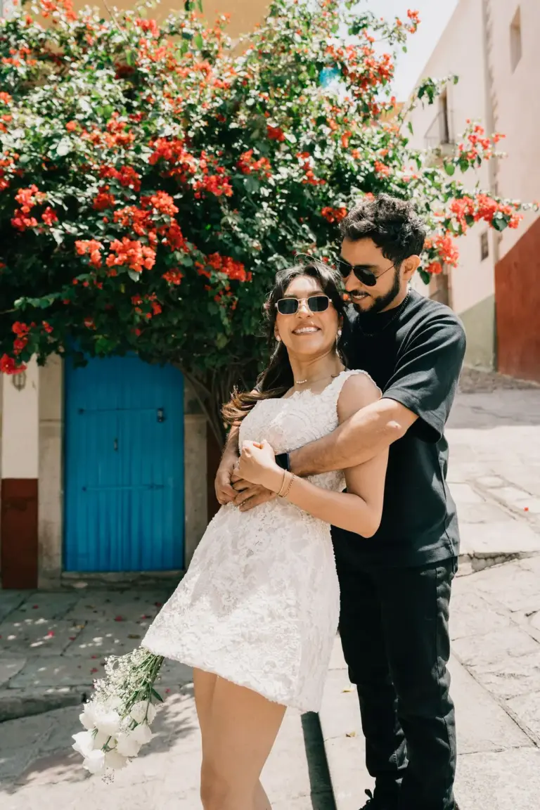 Ella radiante mientras su pareja la abraza bajo la bugambilia roja junto a una puerta azul en Guanajuato