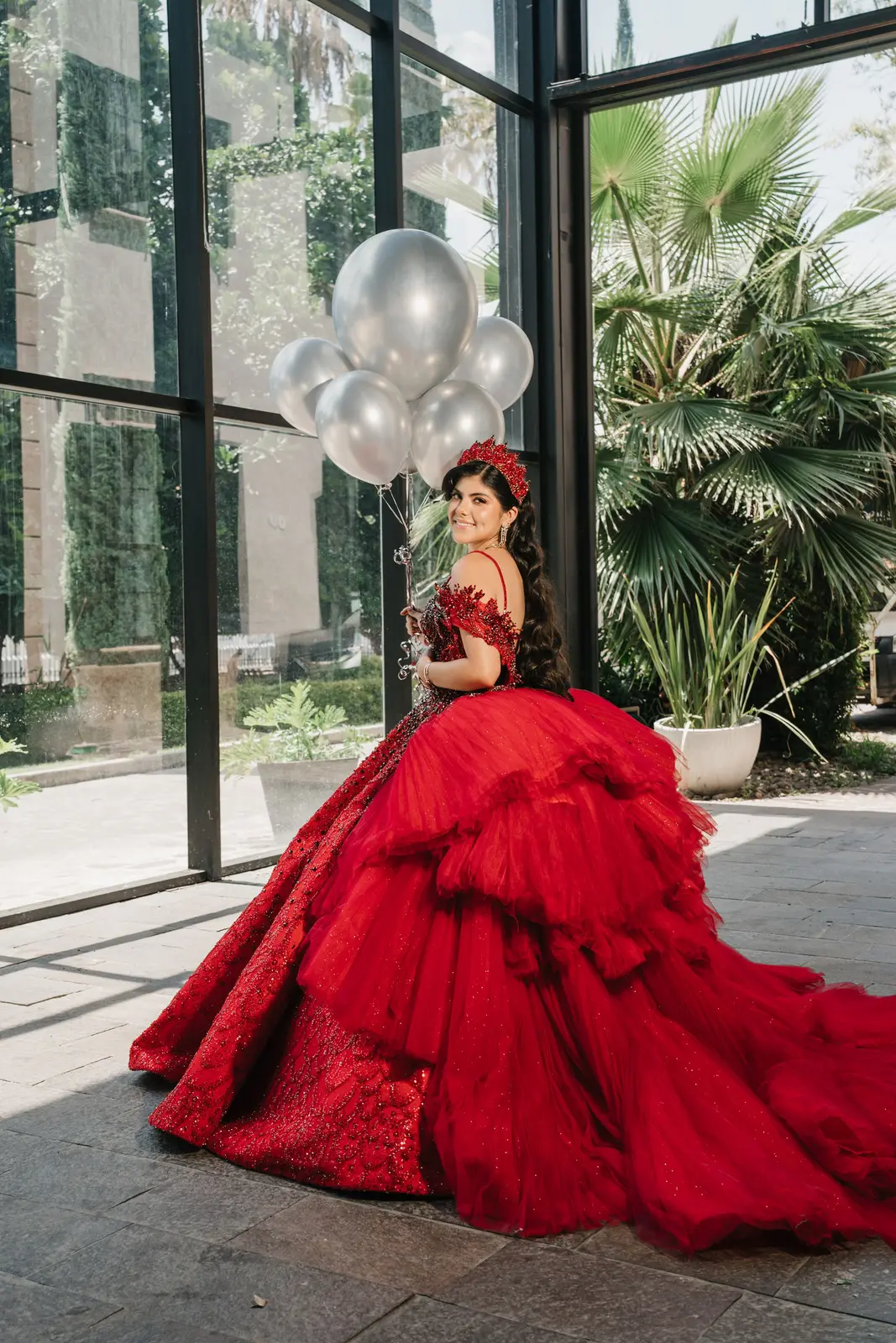 Quinceañera con vestido rojo y globos plateados en un jardín de cristal